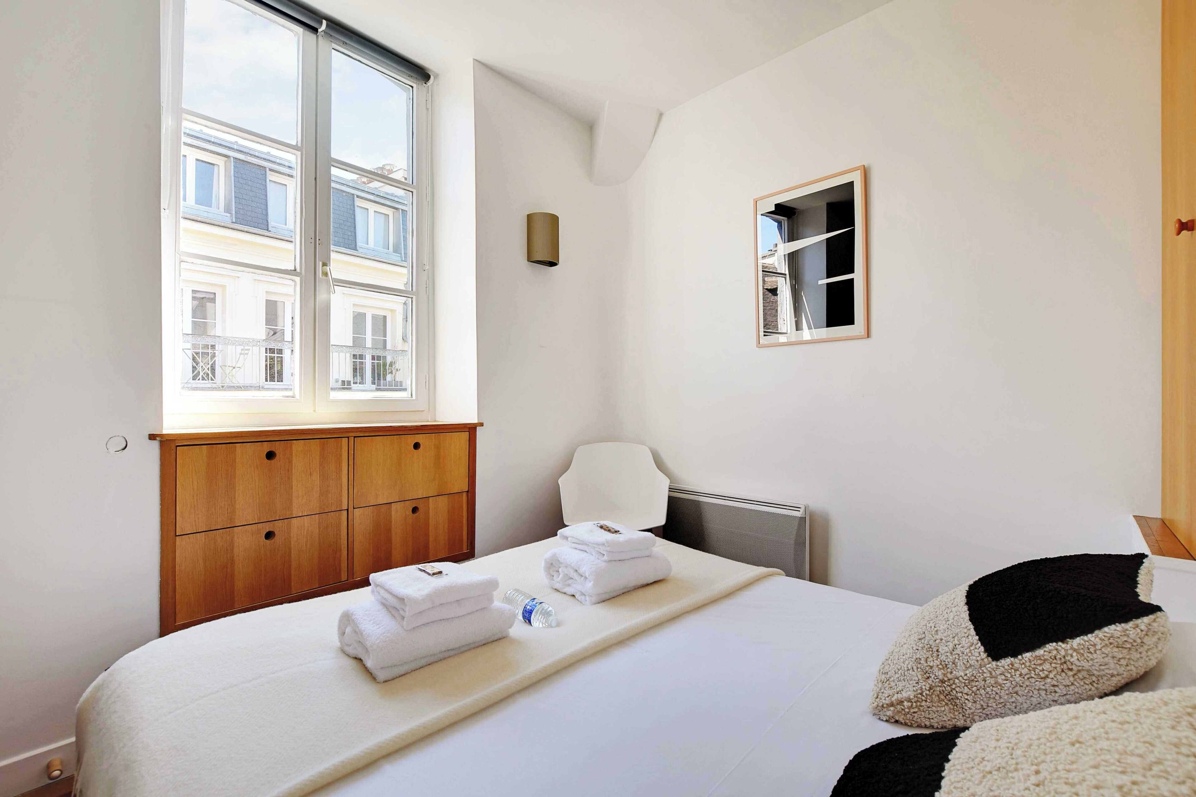 A hotel room with a bed, a wooden wardrobe, a window with a view of buildings, a small mirror, and a towel rack.
