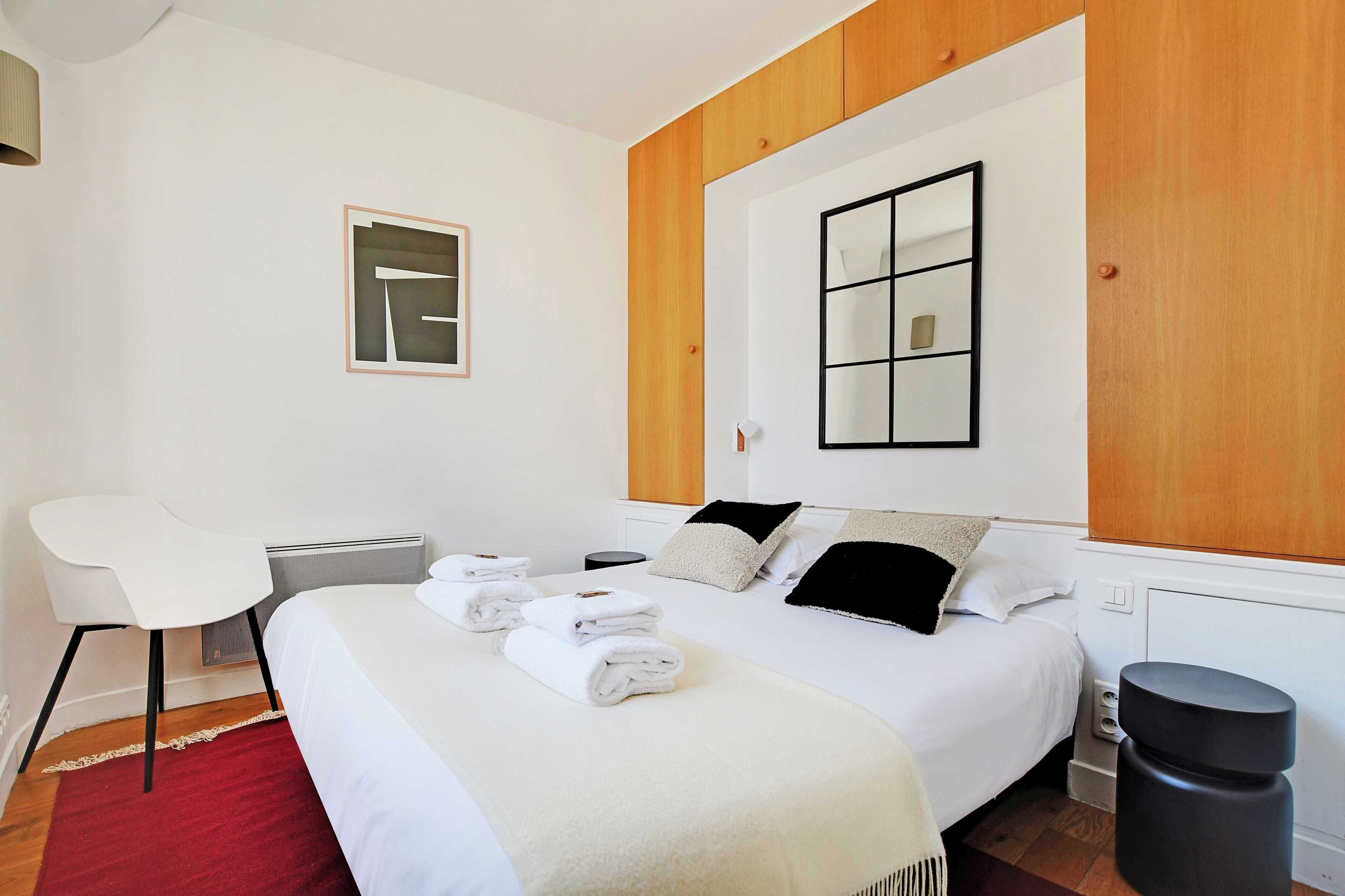 A neatly made bed with white linens and a red rug, a white chair, a small bedside table, and a black trash bin.