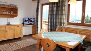 Une pièce chaleureuse avec une table en bois, des chaises et un banc. On y trouve une télévision posée sur une étagère, une fenêtre avec des rideaux et une vue sur l’extérieur.