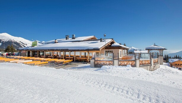 Un bâtiment enneigé doté d’une terrasse et d’un espace couvert, le tout entouré de montagnes.