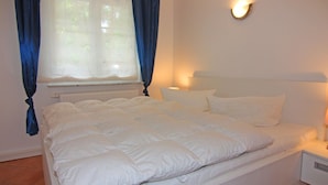 A neatly made bed in a room with a window, a wall-mounted light, and a radiator. - Ferienwohnung in Trassenheide (Trassenheide)