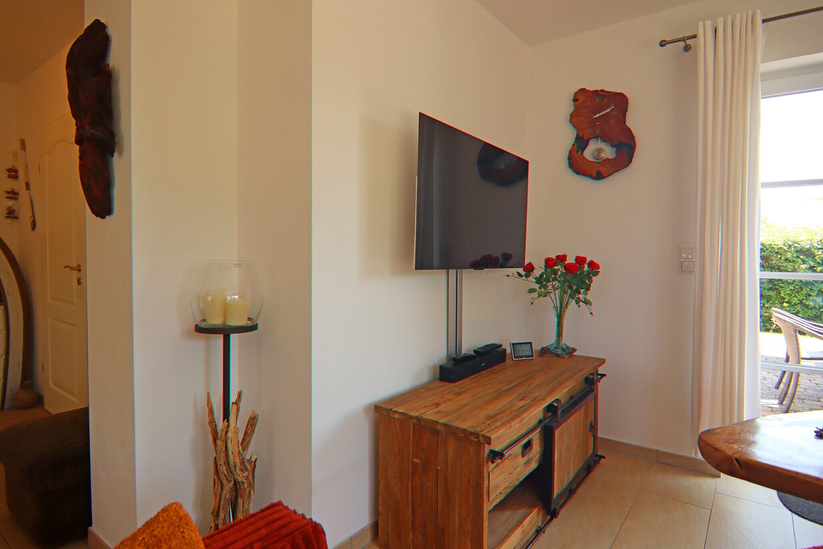 A living room with a wooden coffee table, a flat-screen TV mounted on the wall, and a vase with red flowers on a wooden console table.