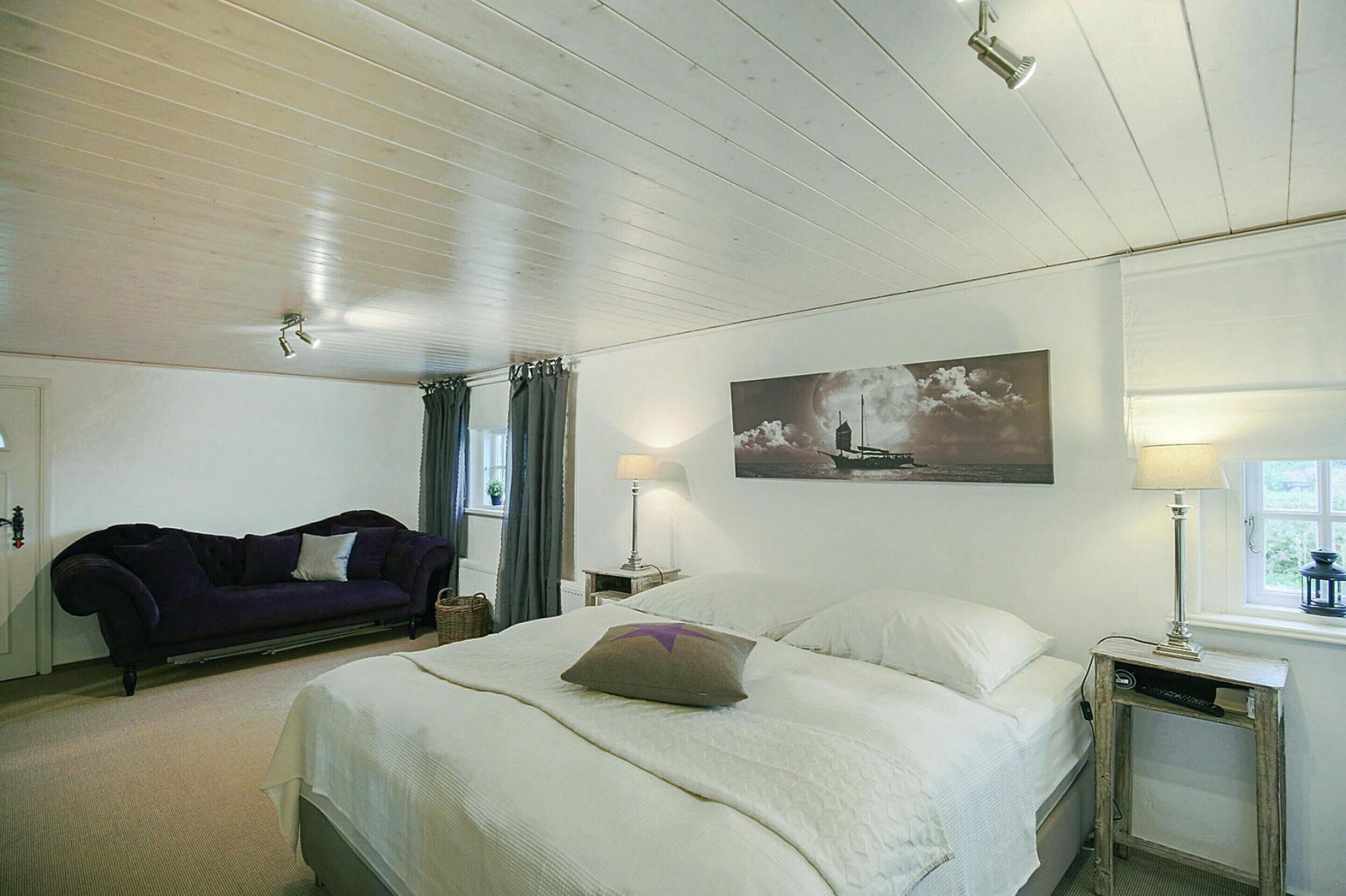 Ein Schlafzimmer mit einem Bett, einem Nachttisch, einer Lampe, einem Fenster, einem Bild an der Wand und einem violetten Sofa.