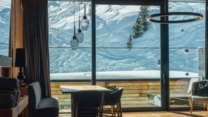 A modern interior with a large window offering a snowy mountain view. There is a wooden dining table with chairs, a sofa, and a lamp. - Apartment in Kaltenbach With Ski-in Access (Kaltenbach)