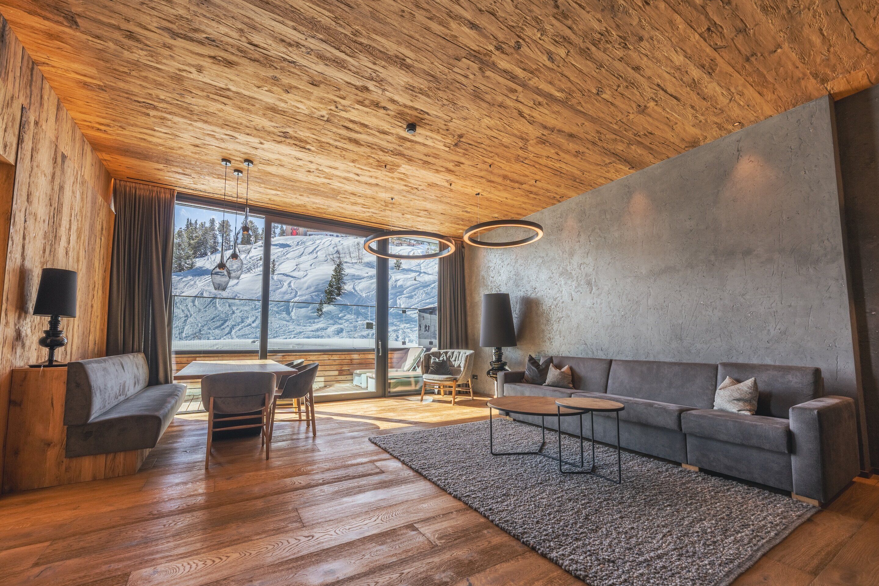 Ein modernes Wohnzimmer mit Holzboden, großen Fenstern, einem grauen Sofa, einem Couchtisch und einem Teppich.