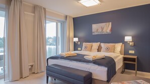 A hotel room with a large bed, a bedside table, a bench, a window with curtains, and a painting on the wall. - Dunenresidenz Prora (Binz)