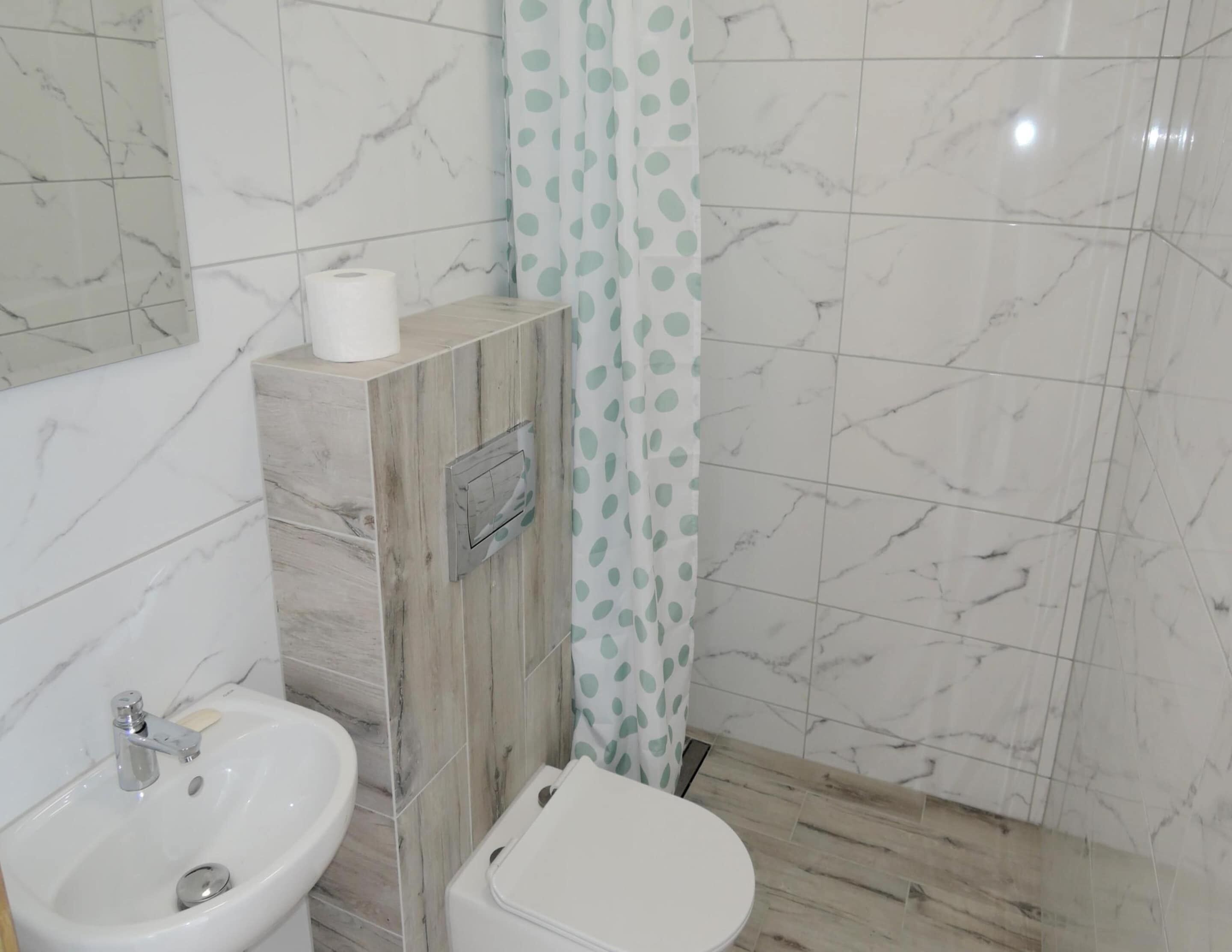A bathroom with a marble-tiled wall, a sink, a toilet, and a shower curtain with a polka dot pattern.