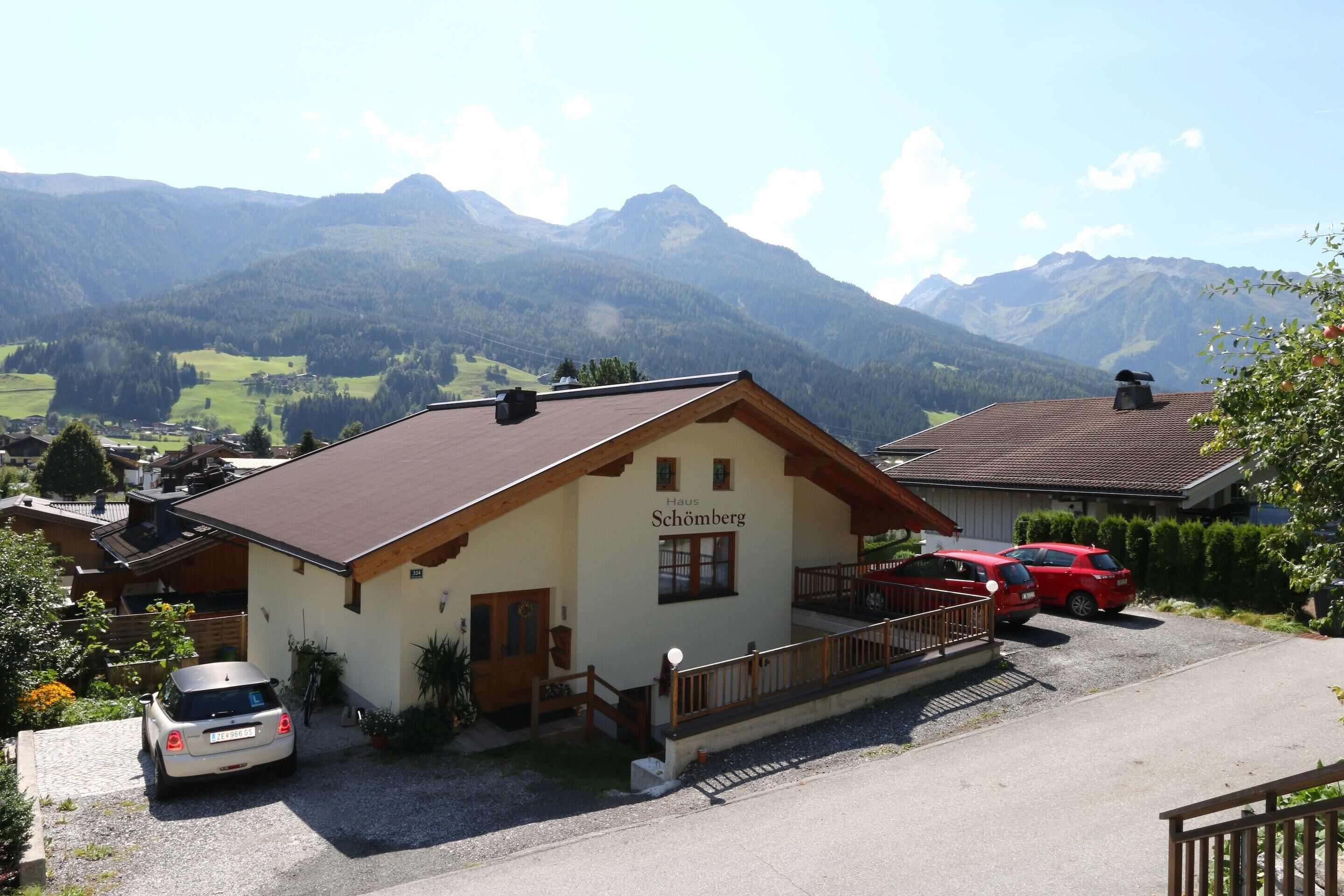 A two-story building with a sign reading "Haus Schömburg" in front. There are two cars parked in front, and a mountain range is visible in the background.
