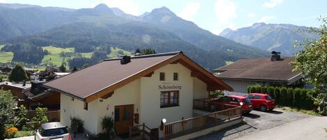 A two-story building with a sign reading "Haus Schömburg" in front. There are two cars parked in front, and a mountain range is visible in the background.