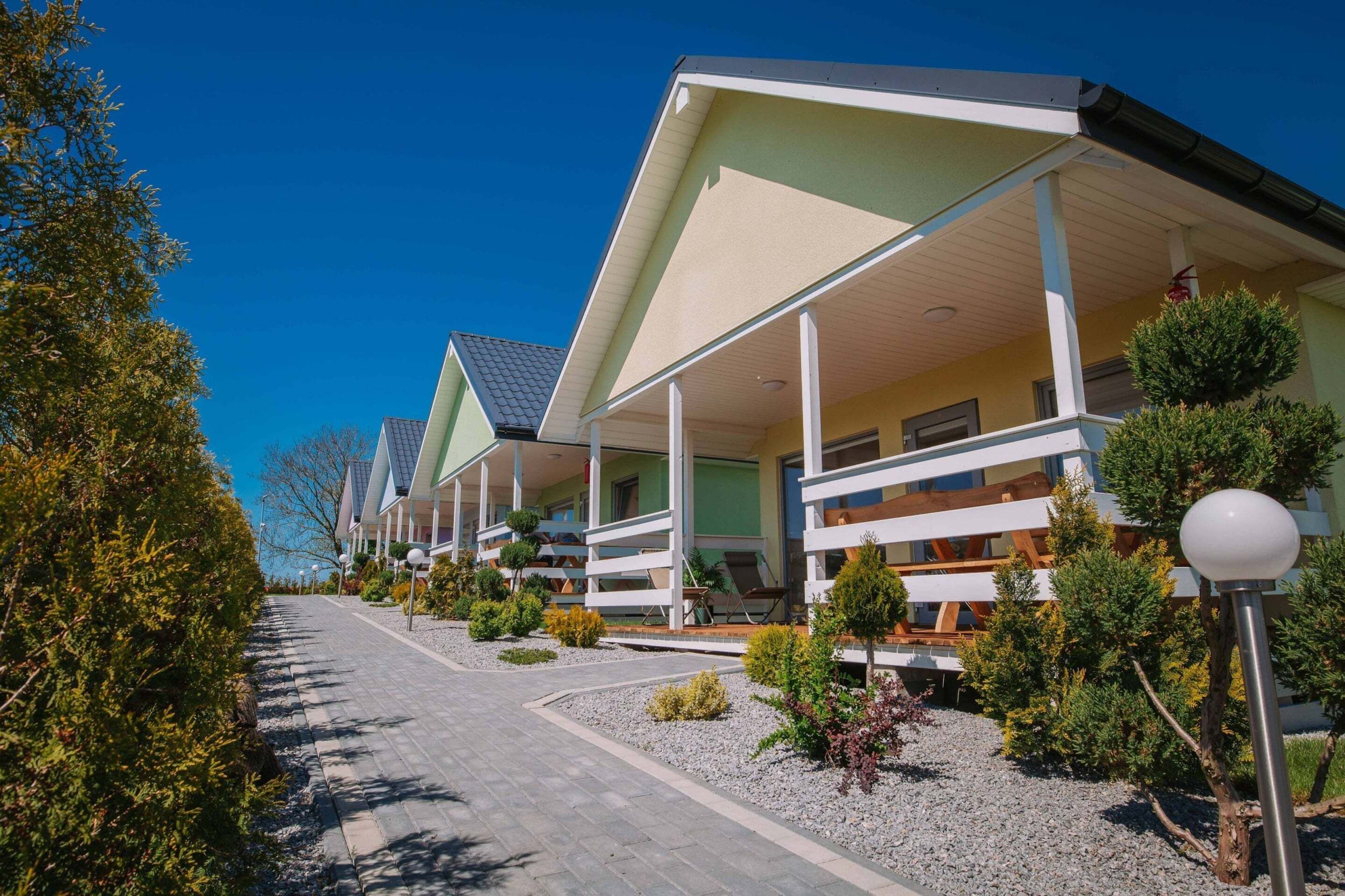 A row of modern, single-story buildings with flat roofs and covered balconies, surrounded by landscaped gardens and a paved walkway.