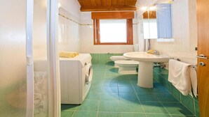 A bathroom with a wooden ceiling, green tiles, a washing machine, two toilets, and a sink with a mirror. - Apartment in Caorle With Heating (Caorle)