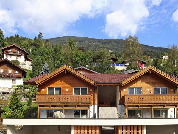 Traditional Alpine-style chalet with a central entrance and balconies on either side, set amidst lush greenery and forested hills.