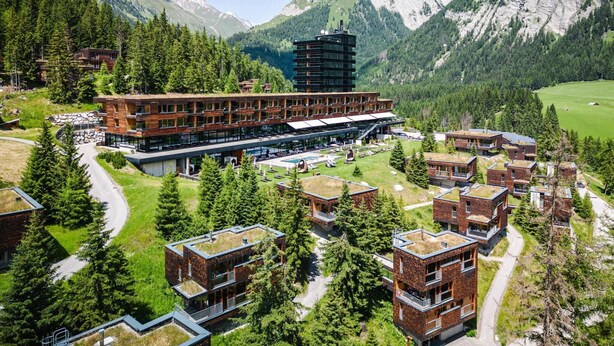 Un moderno complejo hotelero con varias edificaciones, incluyendo una torre destacada, situado en una zona montañosa con abundante vegetación y cielos despejados.