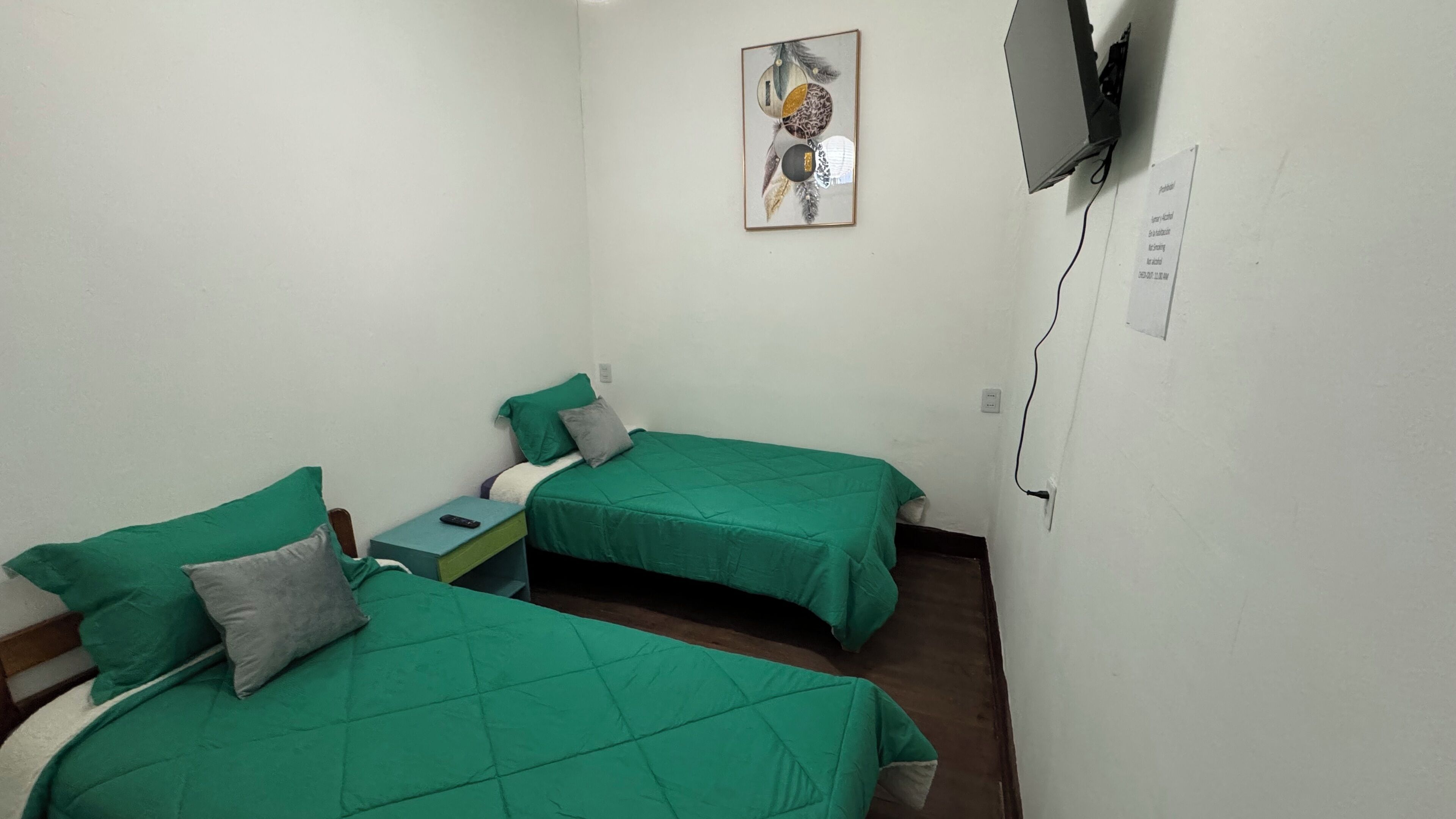 Una habitación pequeña y limpia con dos camas individuales, una mesita de noche con lámpara y televisión instalada en la pared.