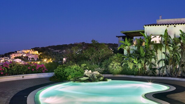 A swimming pool with a wavy edge, surrounded by lush greenery and a view of a hillside with buildings. - Stay in Porto Cervo With Garden Setting (Arzachena)