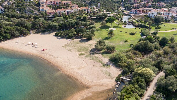 An aerial view of a coastal area with a sandy beach, clear waters, and a cluster of buildings with balconies overlooking the beach. - Stay in Porto Cervo With Garden Setting (Arzachena)