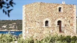 A stone building with arched windows and a balcony, surrounded by greenery and overlooking a body of water. - Stay in Porto Cervo With Garden Setting (Arzachena)