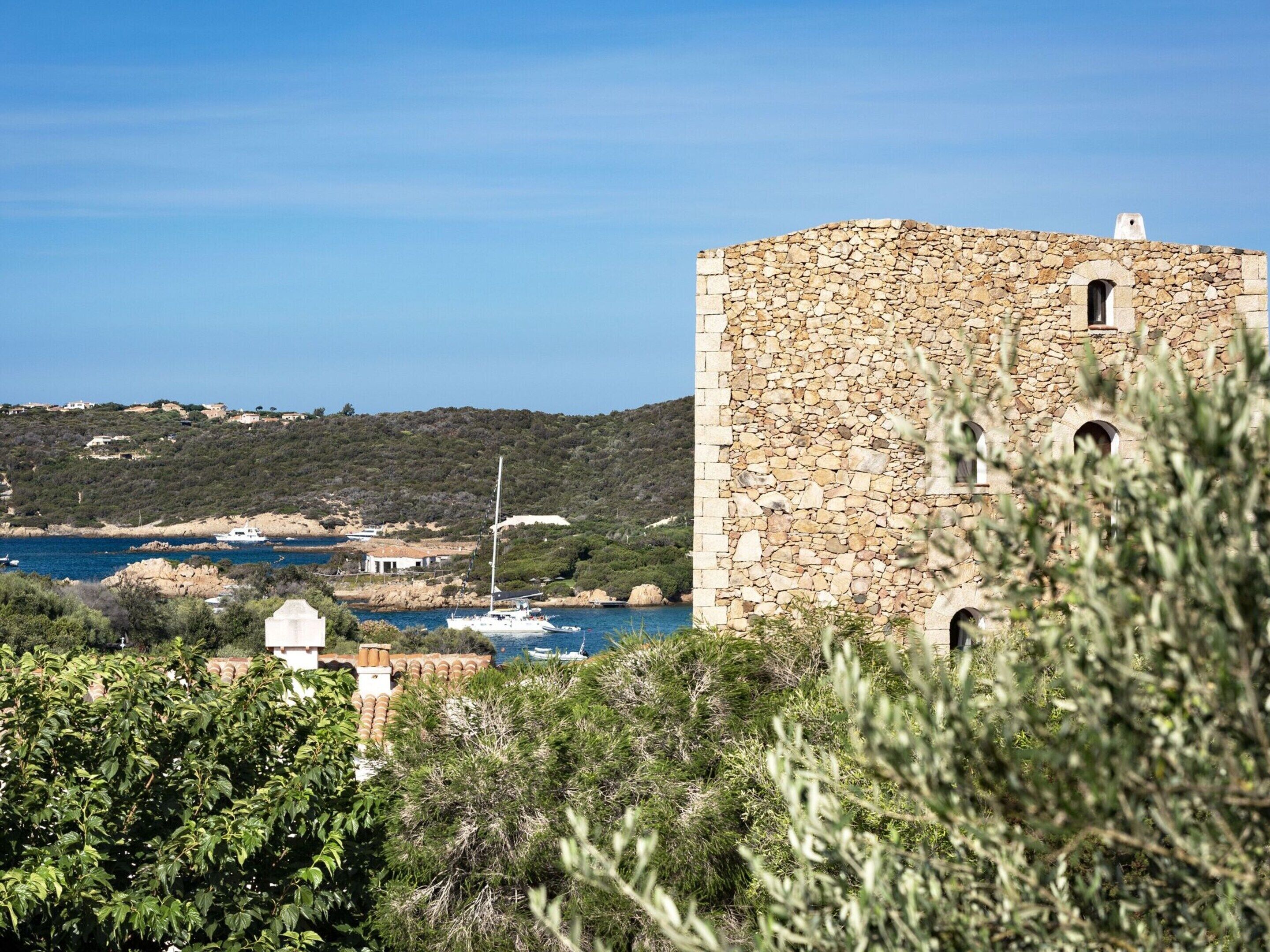 A stone building with arched windows, surrounded by greenery and overlooking a coastal area with boats.