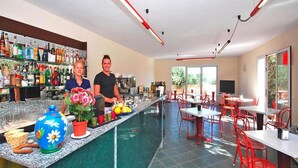 Un espace bar avec un comptoir, une variété de bouteilles et un grand vase décoratif. La salle à manger est aménagée avec des tables et des chaises, et une grande fenêtre laisse entrer la lumière naturelle.