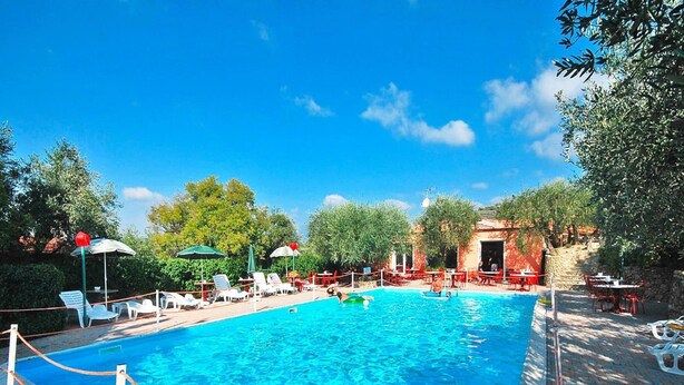 Une piscine entourée d’arbres et d’un bâtiment en pierre, avec des chaises longues et des parasols à proximité.