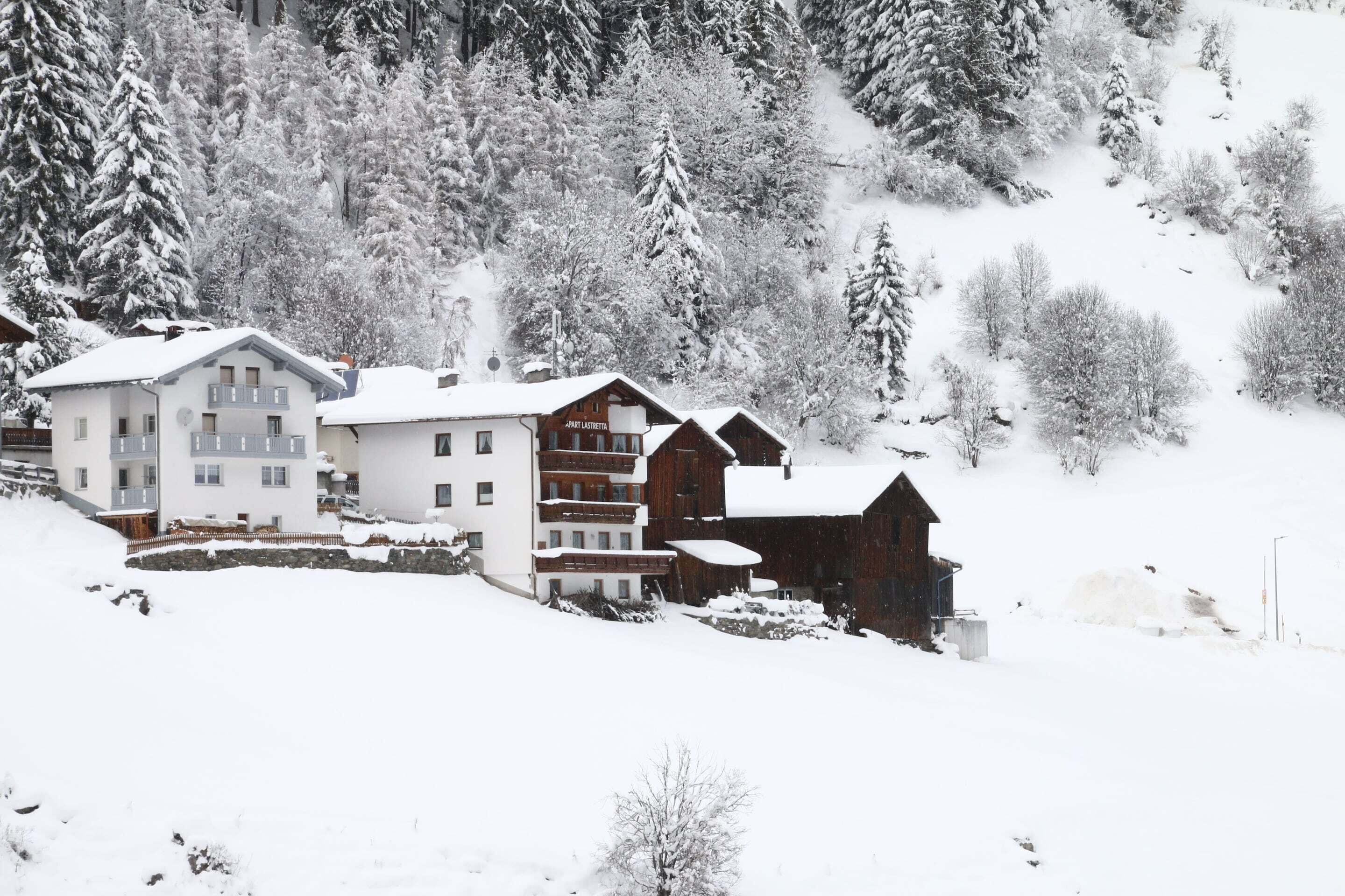 A snow-covered mountain village with multi-story buildings, including a hotel with balconies and a wooden cabin.