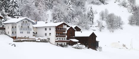 A snow-covered mountain village with multi-story buildings, including a hotel with balconies and a wooden cabin.