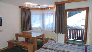 Ett hotellrum med ett skrivbord i trä, två sängar med rutigt sänglinne, utsikt över ett snötäckt berg genom en balkongdörr och ett fönster med tunna, genomskinliga gardiner.