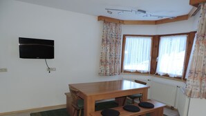 Ett rum med ett matbord i trä, bänkar, en tv som är monterad på väggen och två fönster med gardiner.