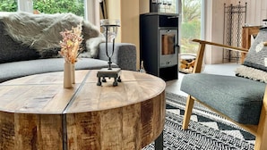 A cozy living room with a sofa, a round wooden coffee table, a fireplace, and a view of greenery outside. - Holiday Home Seeblick With Sauna on Lake Dummer, Dummer (Dümmer)