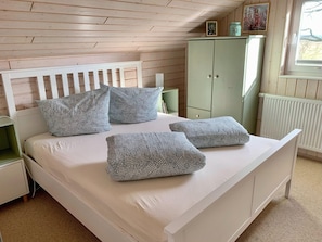 Une chambre avec un lit blanc, des coussins à motifs, une armoire verte et une fenêtre donnant sur un beau paysage.