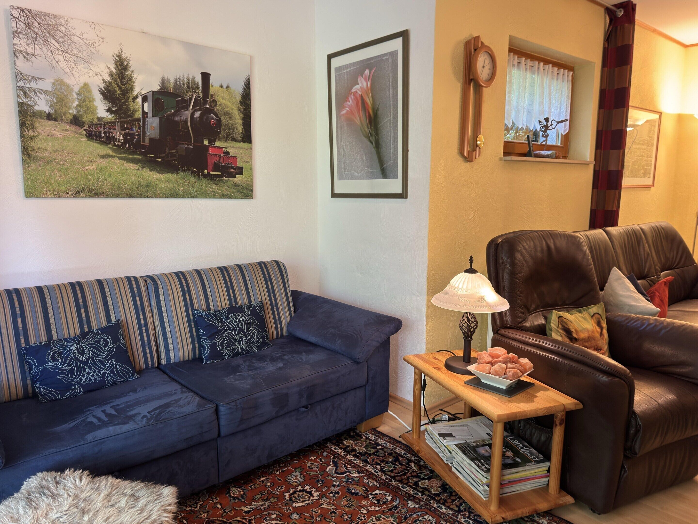 A living room with a dark blue sofa, a wooden coffee table with magazines, a brown leather armchair, and a painting of a train on the wall.