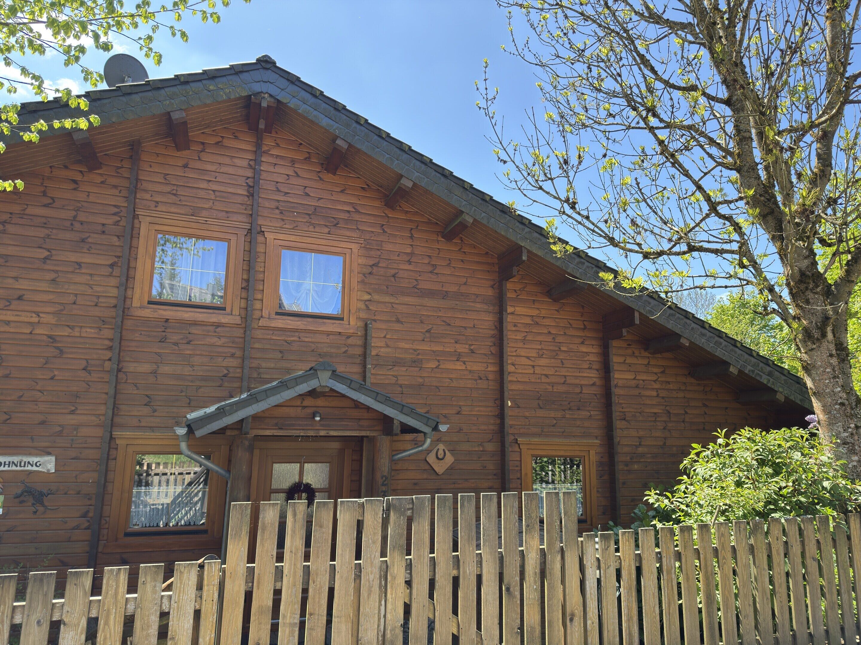 A wooden cabin with a sloped roof, two windows, and a wooden fence in front.