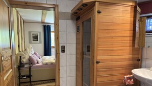 Eine Holzsauna mit Glastür, ein Bett mit blumigem Bettbezug sowie ein Badezimmer mit Waschbecken und Handtuchhalter.