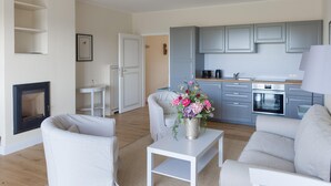 A modern living room with a fireplace, white sofas, a coffee table with a vase of flowers, and a kitchen with grey cabinets and a stove. - Ferienwohnung mit Kamin im Gut Ulrichshusen (Moltzow)