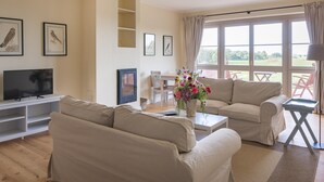 A living room with beige sofas, a flat-screen TV, a coffee table with a vase of flowers, and a large window with curtains. - Ferienwohnung mit Kamin im Gut Ulrichshusen (Moltzow)