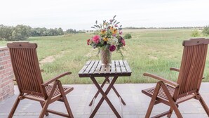A patio with two wooden chairs and a table, a vase of flowers, and a view of a grassy field. - Ferienwohnung mit Kamin im Gut Ulrichshusen (Moltzow)