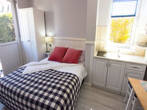 Une chambre avec une couvre-lit à carreaux, une table de chevet avec une lampe, une fenêtre donnant sur les arbres et une kitchenette équipée d’armoires et d’un évier.