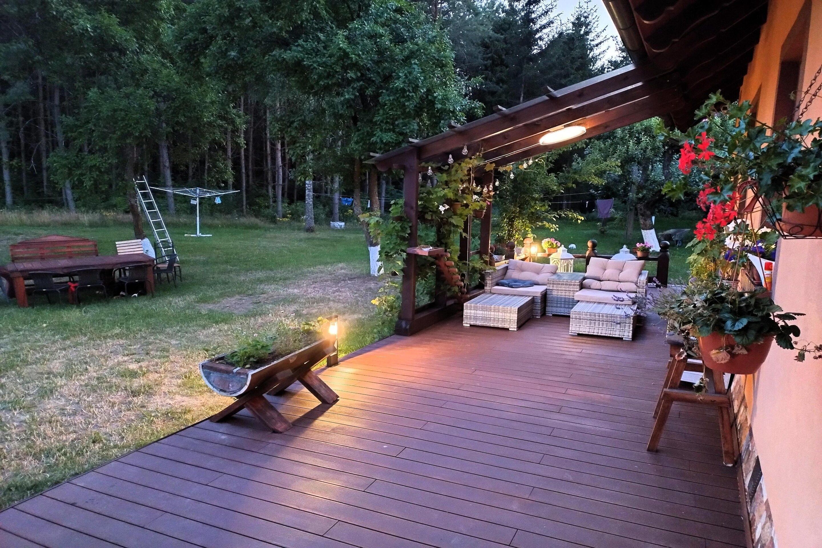 Une terrasse en bois avec des meubles d’extérieur, une pergola illuminée de guirlandes lumineuses et un espace jardin agrémenté de plantes en pot.