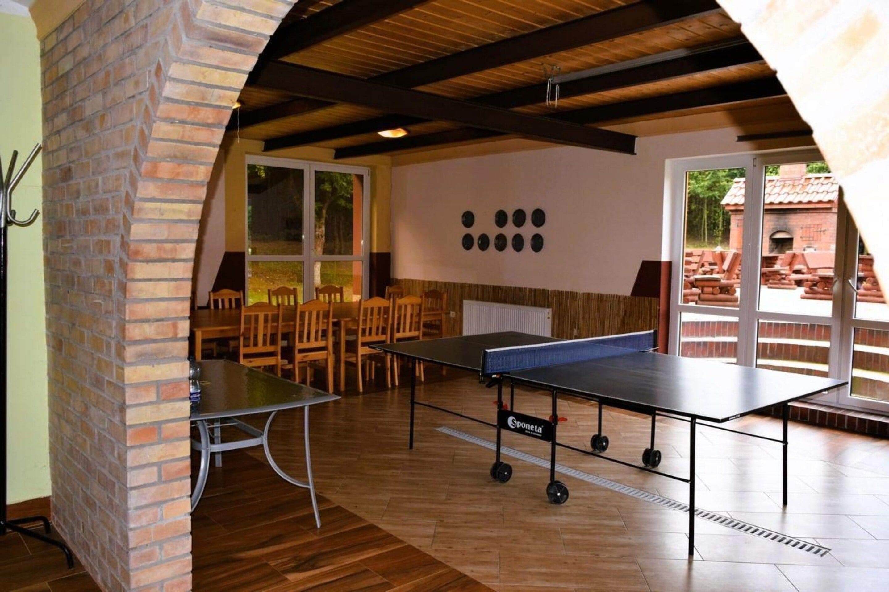 A room with a ping pong table, wooden chairs, a brick wall, and a large window.