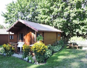 Une cabane en bois avec un porche, entourée d’un jardin où poussent des fleurs jaunes et où se trouve une table de pique-nique.