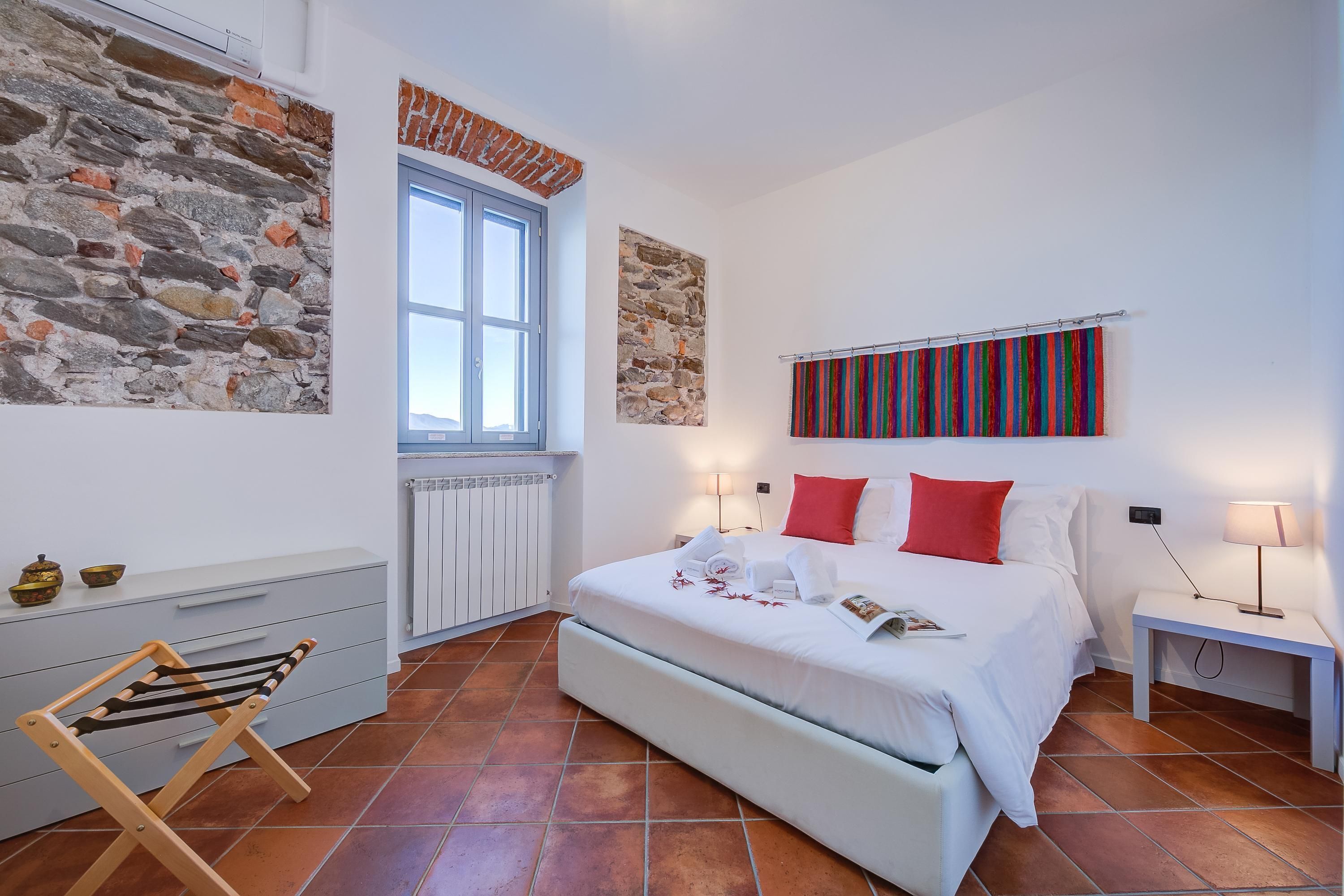 Quarto com parede de pedra, janela, cama com roupa de cama branca e almofadas vermelhas, mesa de cabeceira com abajur e uma cadeira de madeira pequena.