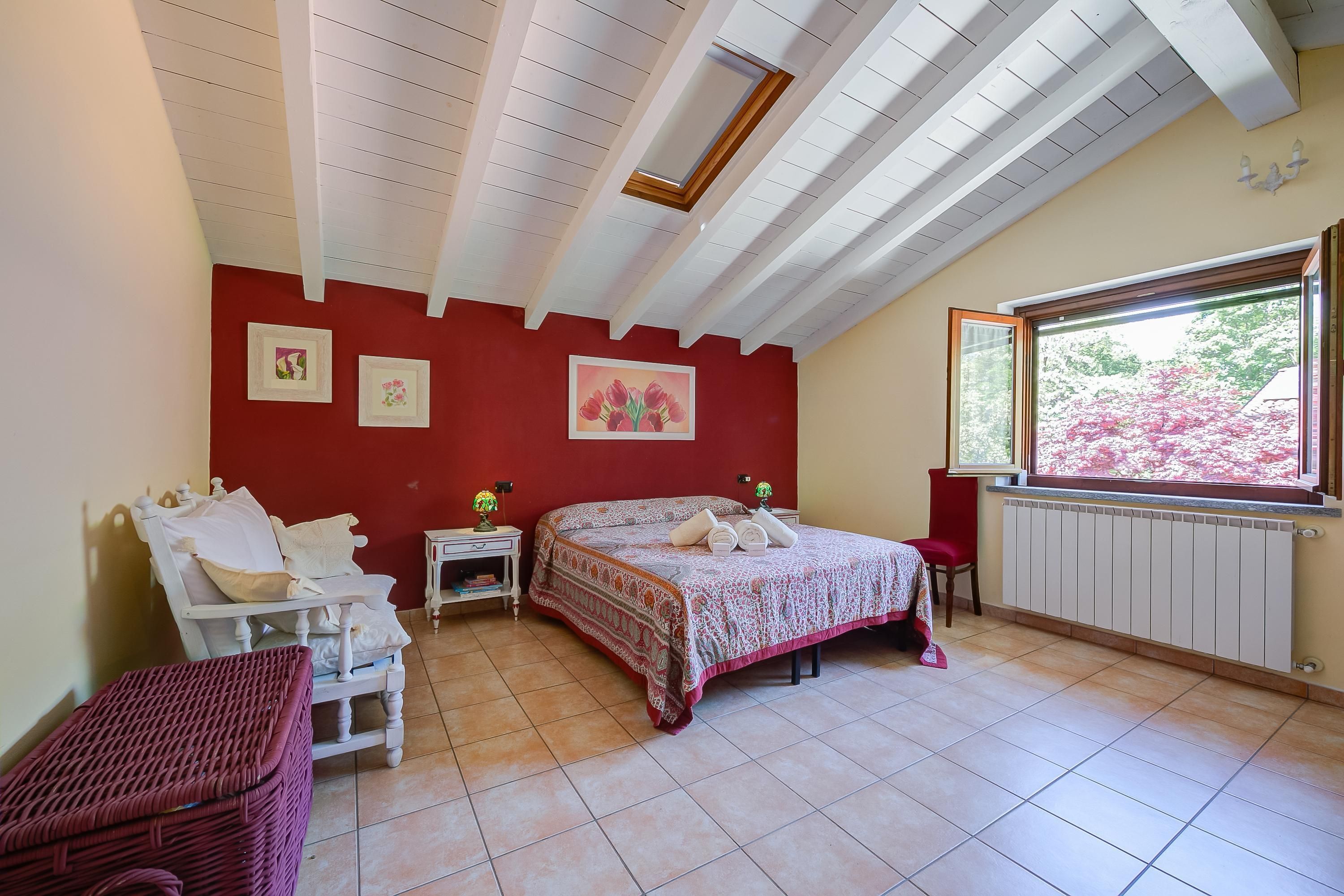 A bedroom with a bed, a chair, a small table, a window with a view of trees, and a skylight.
