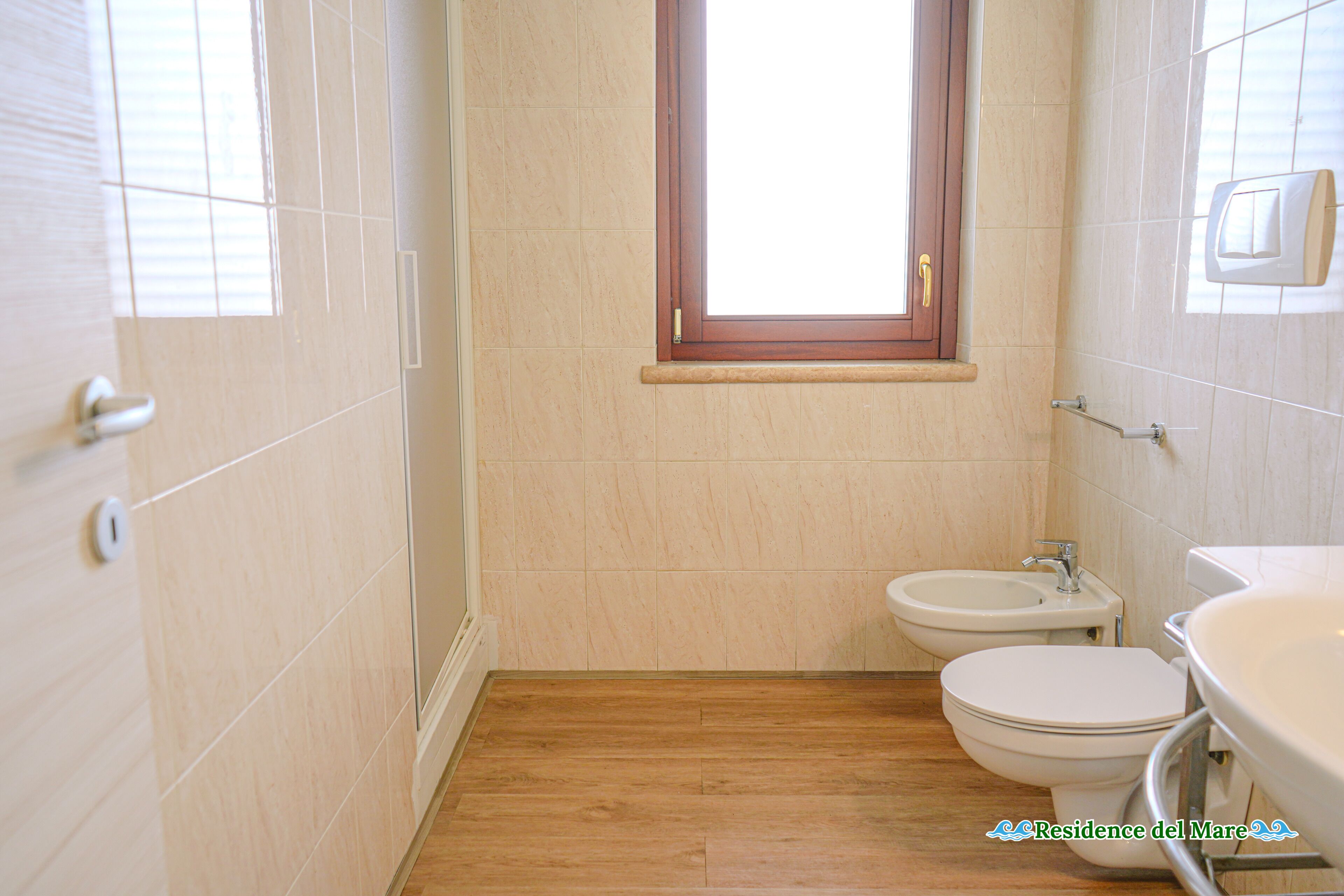A small bathroom with a wooden floor, a window, a toilet, and a bidet.