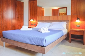 A bedroom with a wooden bed frame, a blue bedspread, two bedside tables with lamps, and wooden wardrobes. - residence del mare (Ardea)