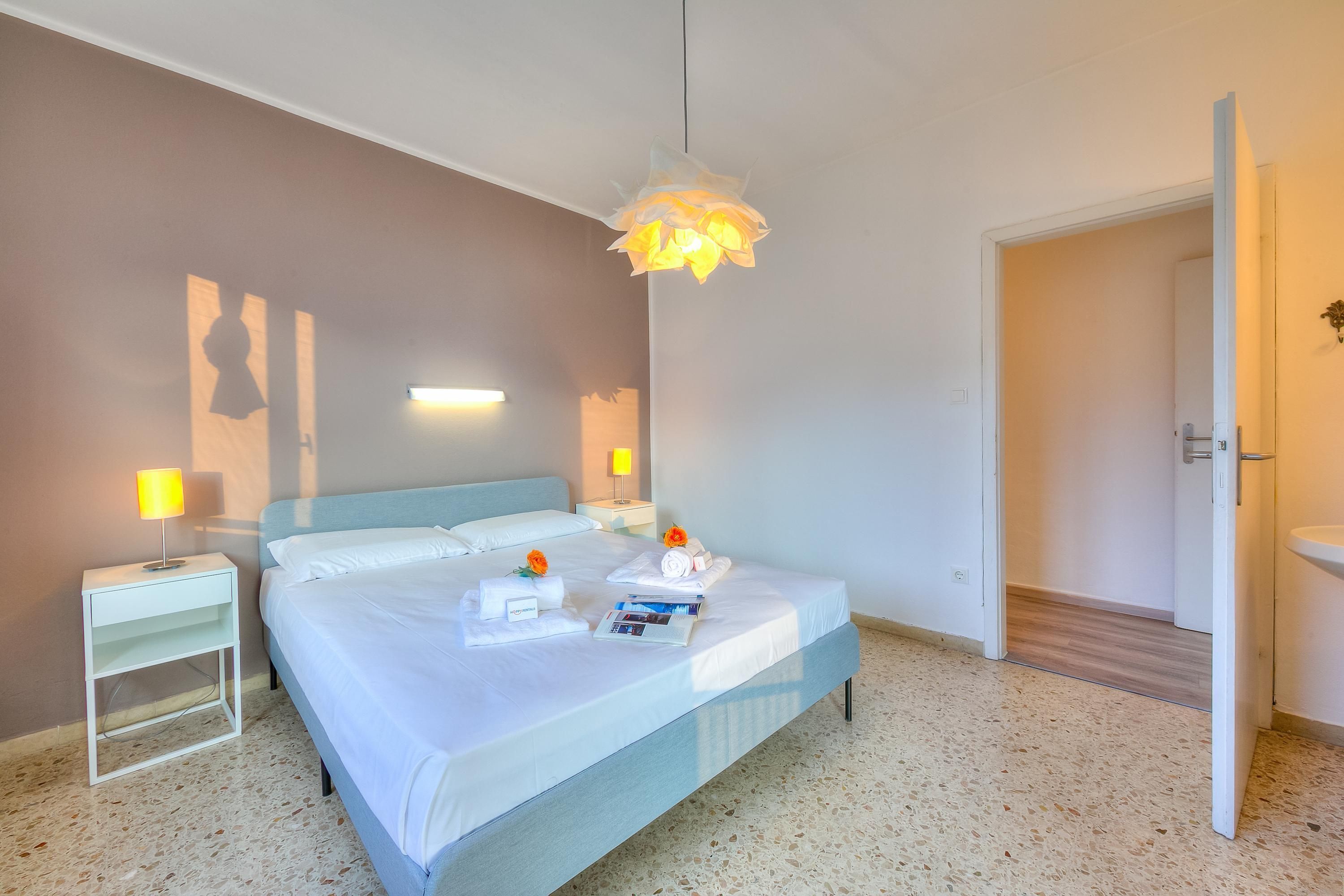 A neatly made bed with a white comforter and pillows, a bedside table with a lamp, and a decorative hanging light fixture.