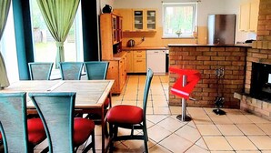 A dining area with a table and chairs, a kitchen with wooden cabinets and a brick fireplace, and a window with curtains. - Holiday Home in Nowe Warpno Jagoda (Nowe Warpno)