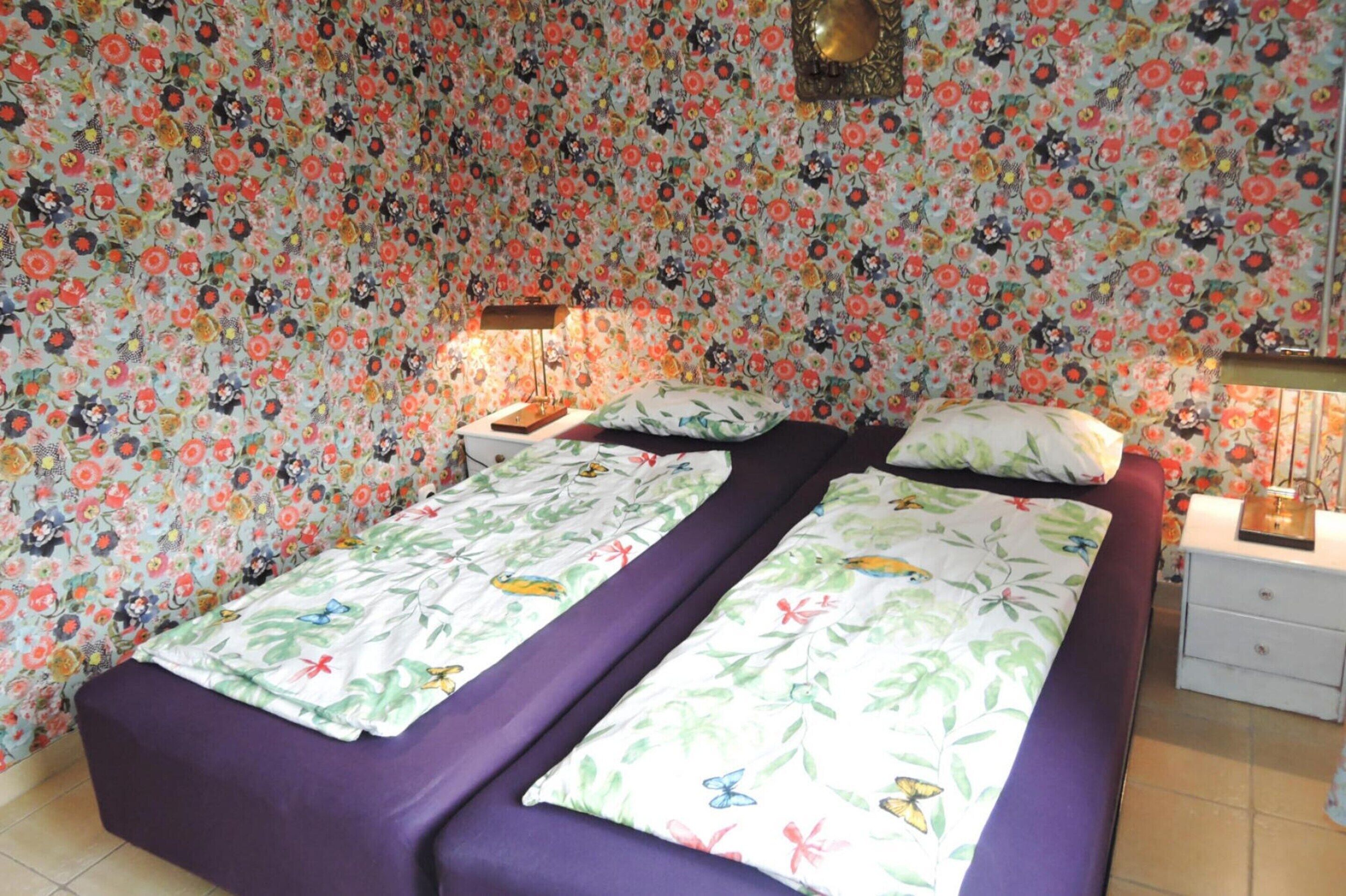 A room with two beds, floral wallpaper, a nightstand with a lamp, and a bedside table with a lamp.