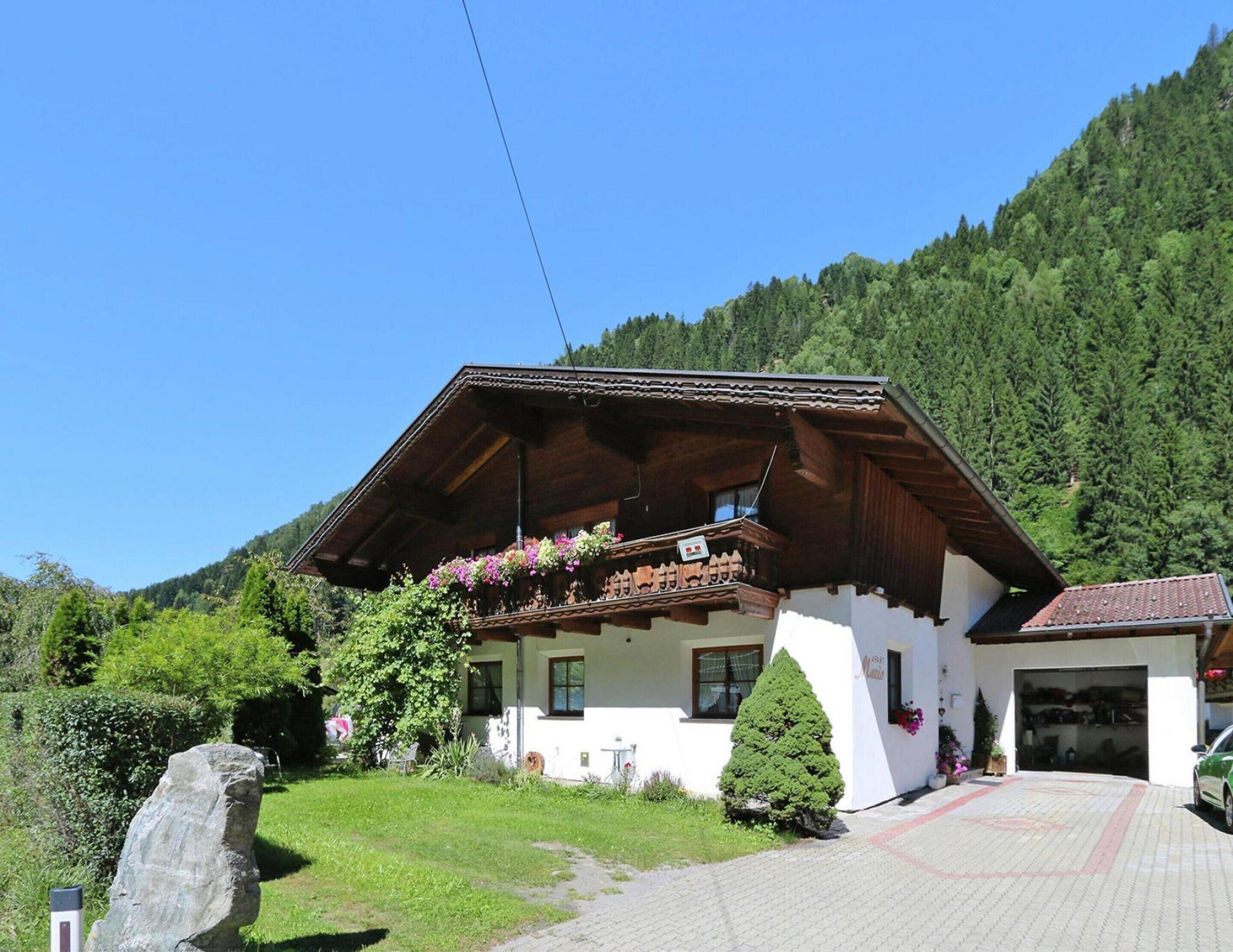 Un chalet traditionnel doté d’un balcon en bois, entouré d’une végétation luxuriante et adossé à un paysage montagneux.