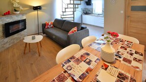 Un salon chaleureux avec une cheminée, un canapé gris, une table à manger agrémentée d’un set de table sur le thème du café, et un escalier menant à l’étage supérieur.
