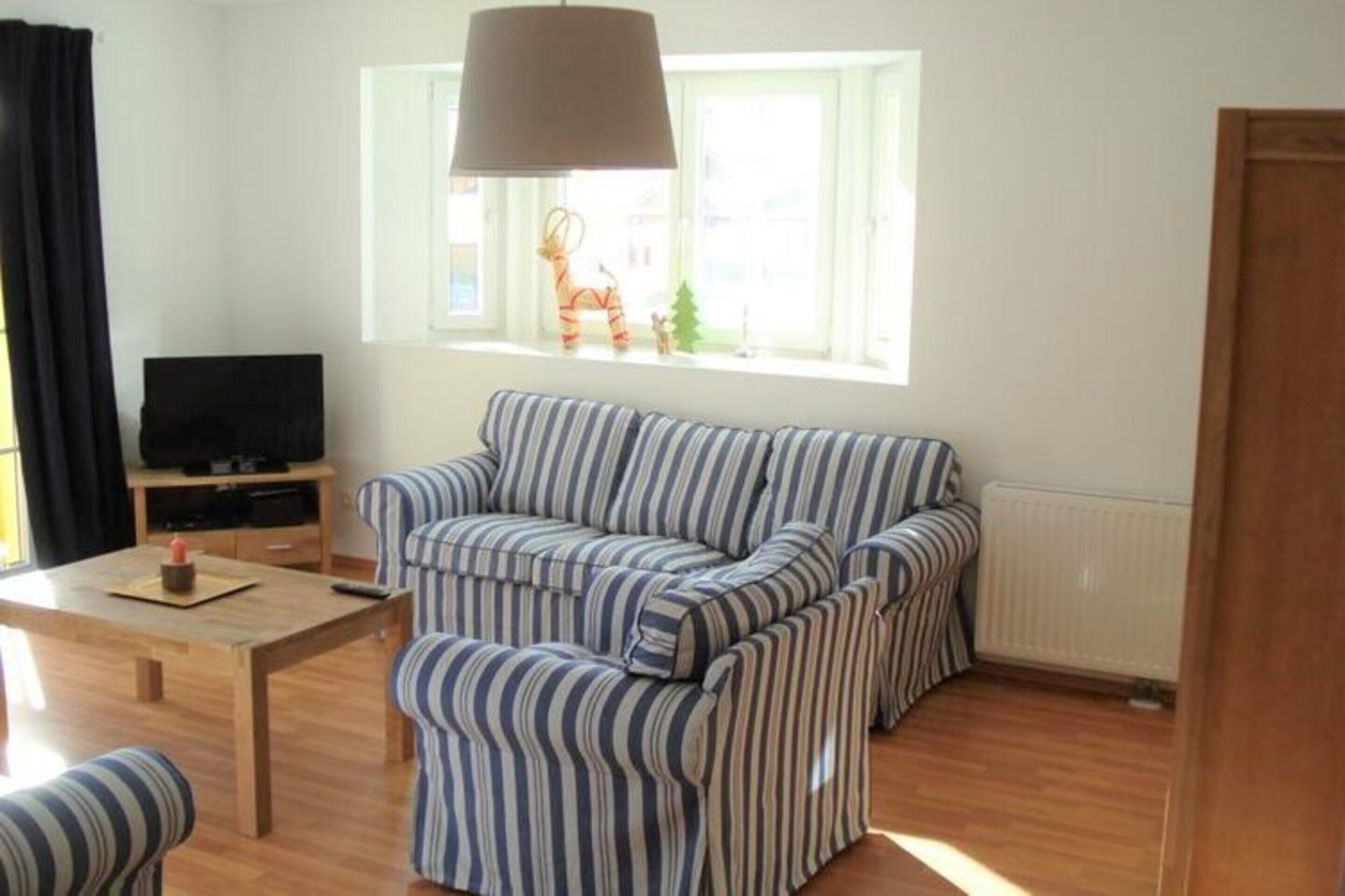 Una sala de estar con un sofá a rayas, una mesa de centro de madera, un televisor sobre un mueble bajo y una ventana con vistas a edificios.